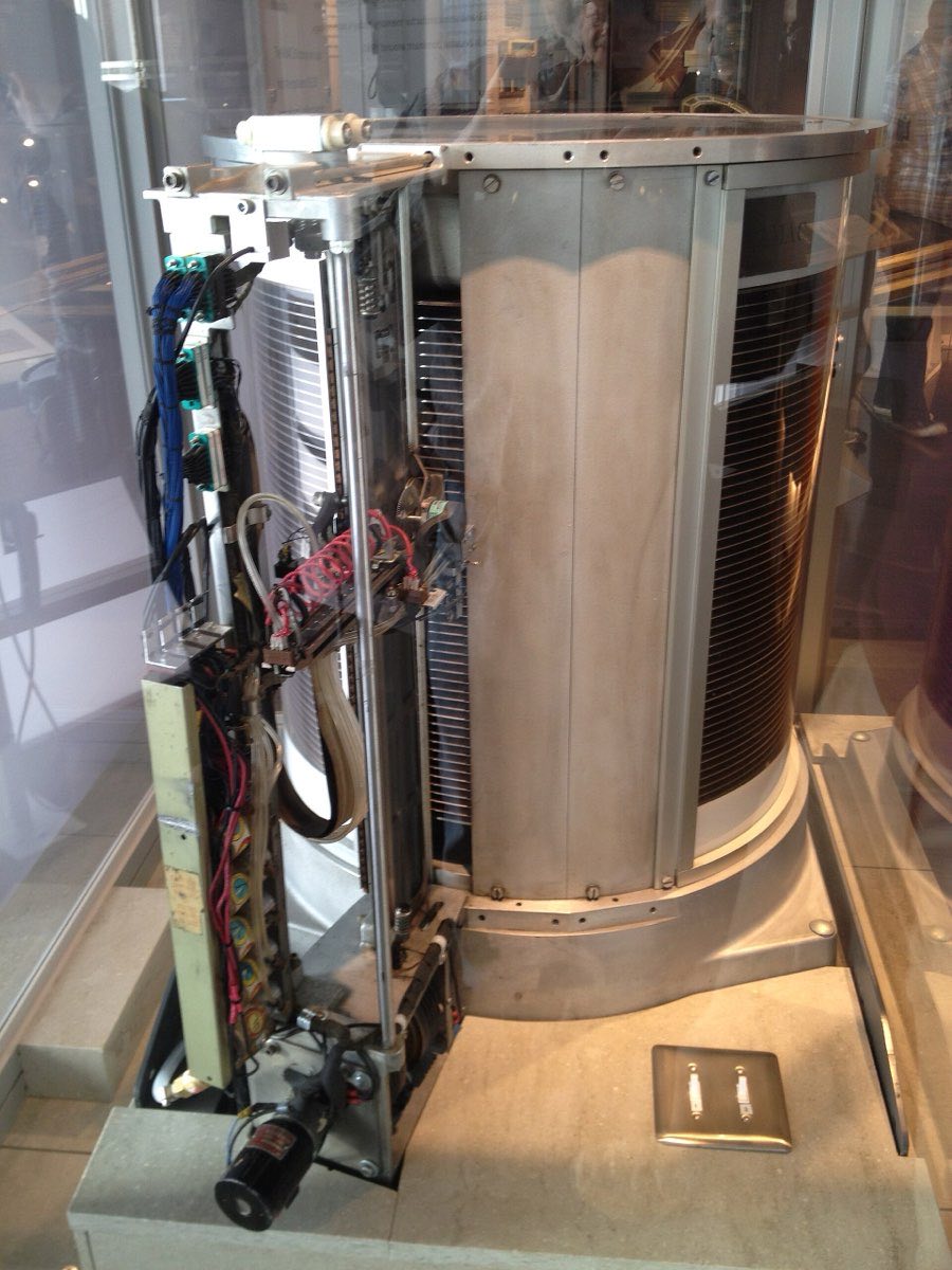 A tall stack of metal disk platters beside its access arm and wiring, behind museum glass.