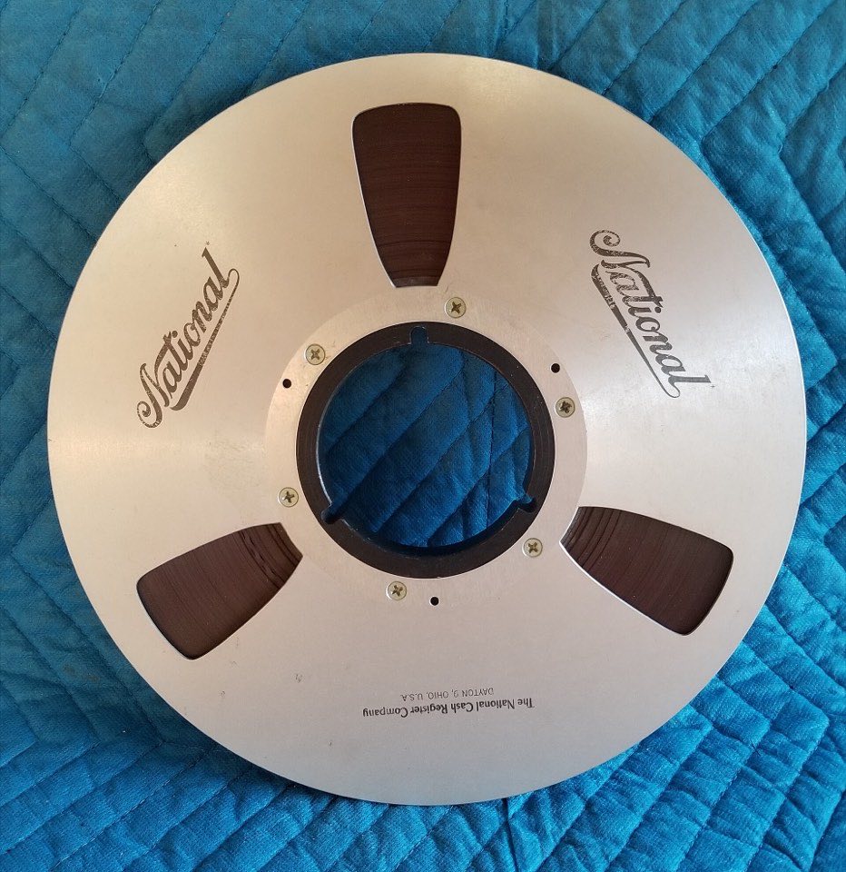A metal computer tape reel with three cut-outs, lying on a quilted cloth.
